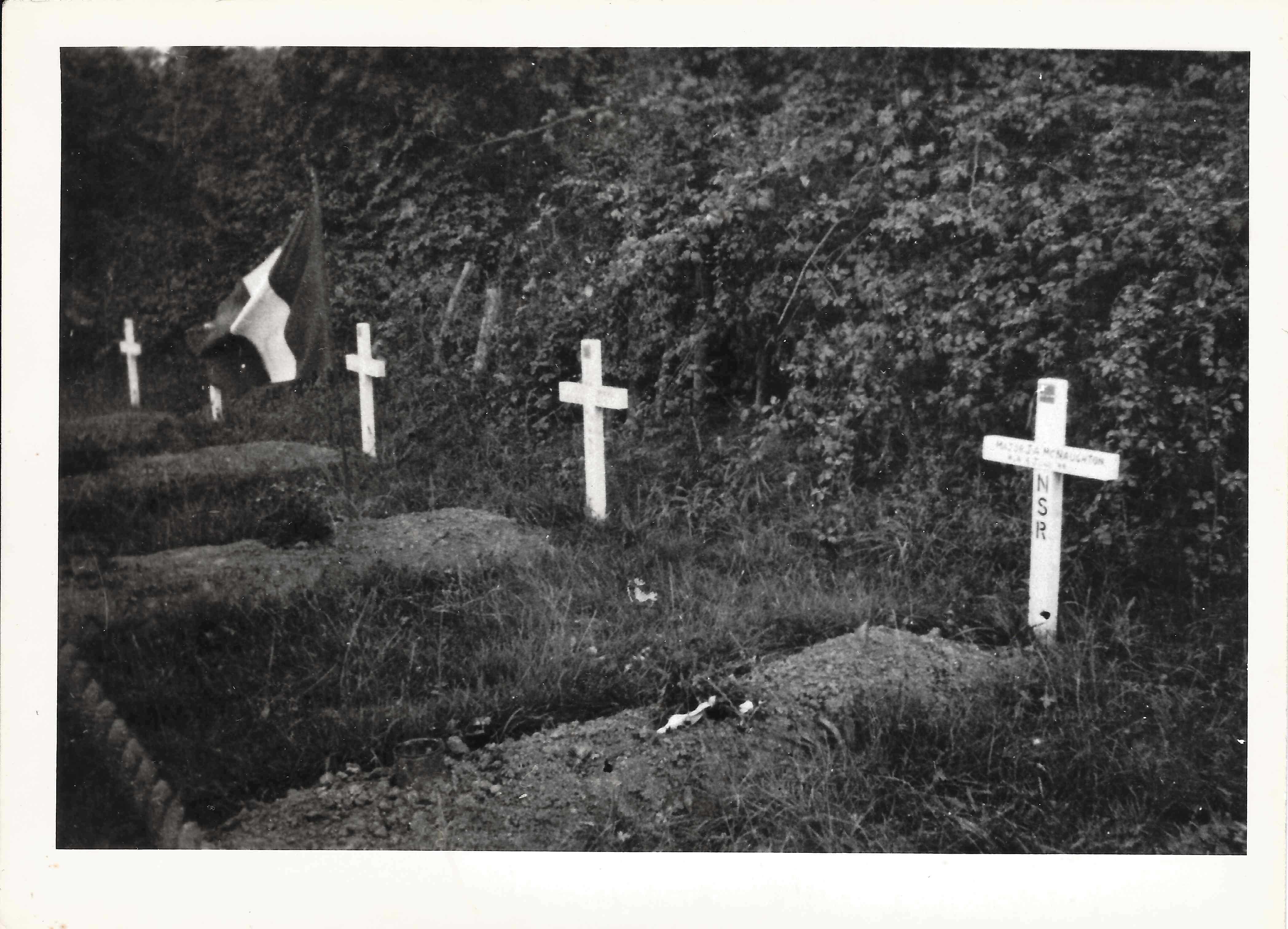 Black and white photograph. A white cross stands at the head of a fresh grave, beside several others and near a French flag. The graves are under several trees. Each has a bouquet on top of it.